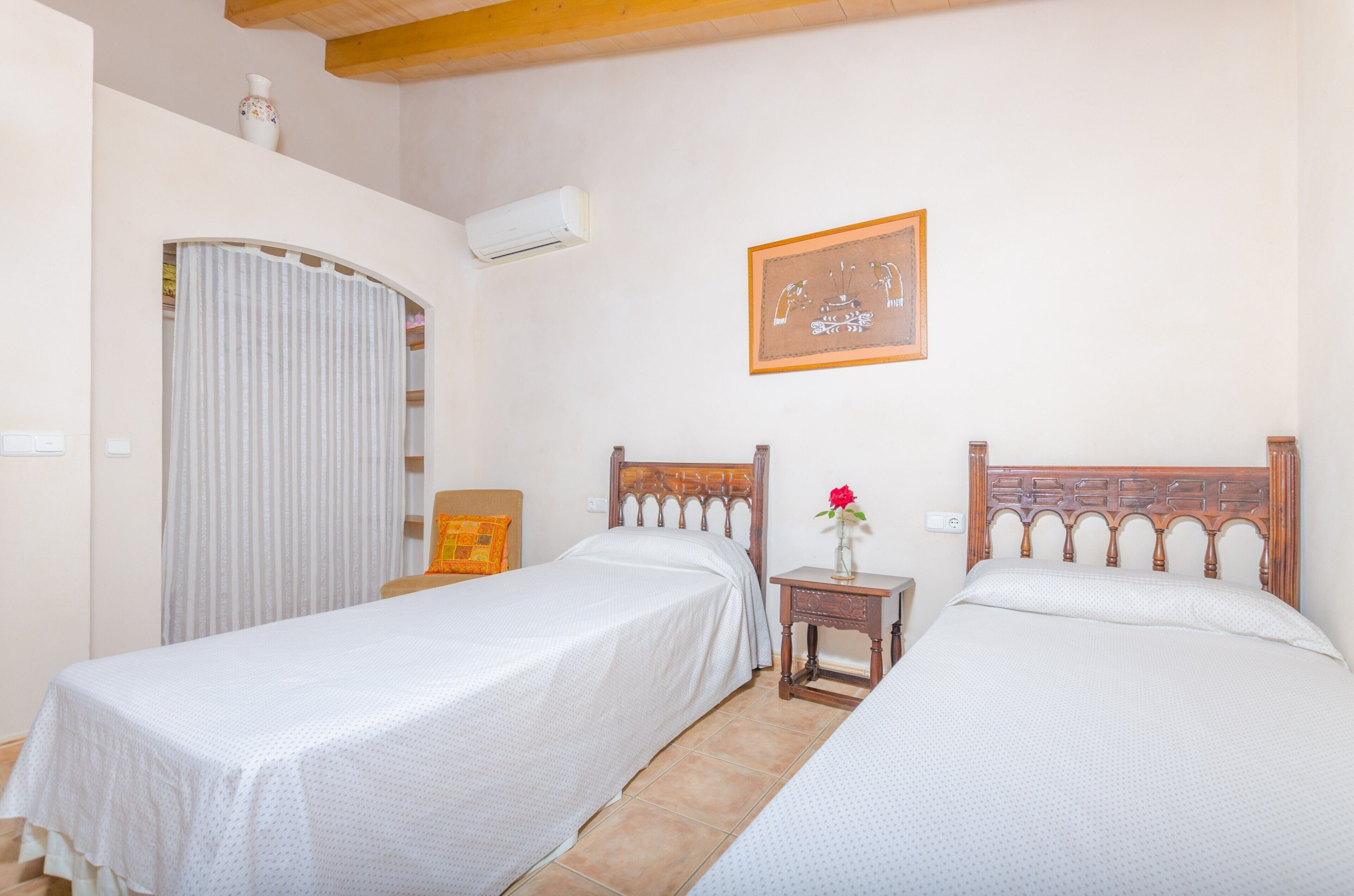 Una habitación con dos camas individuales, una mesita de noche con un florero y un cuadro en la pared, además de un equipo de aire acondicionado.