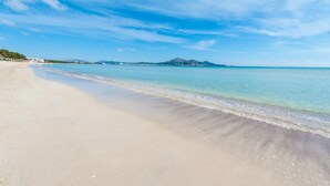 A beach with clear blue water, a sandy shore, and distant mountains. - SA MATA - Spectacular Majorcan finca with private pool enclosed by the trees and greenery (Campanet)