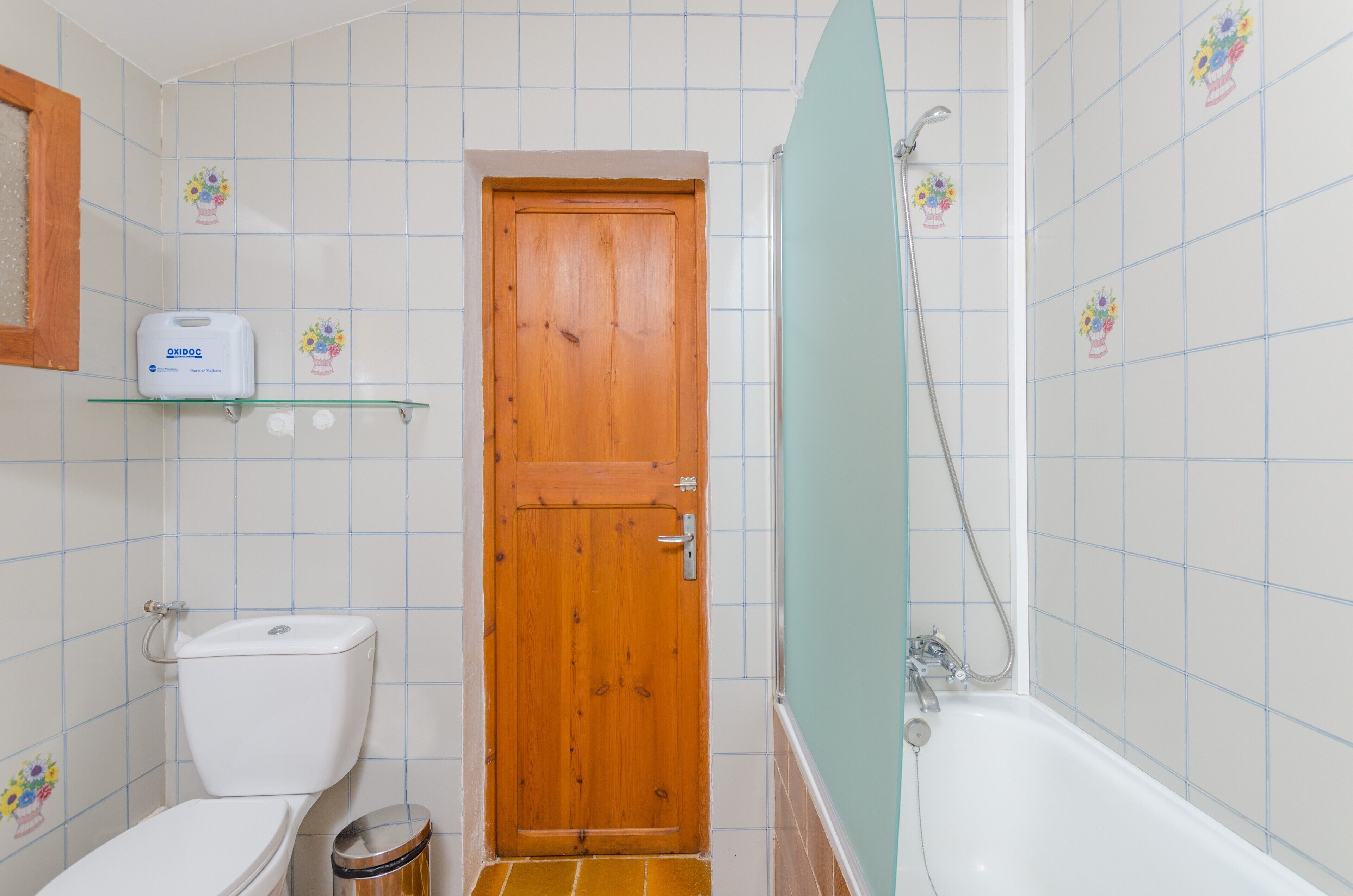 A bathroom with a wooden door, a white toilet, a trash bin, a shelf with a soap dispenser, and a bathtub with a showerhead.