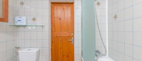 A bathroom with a wooden door, a white toilet, a trash bin, a shelf with a soap dispenser, and a bathtub with a showerhead.