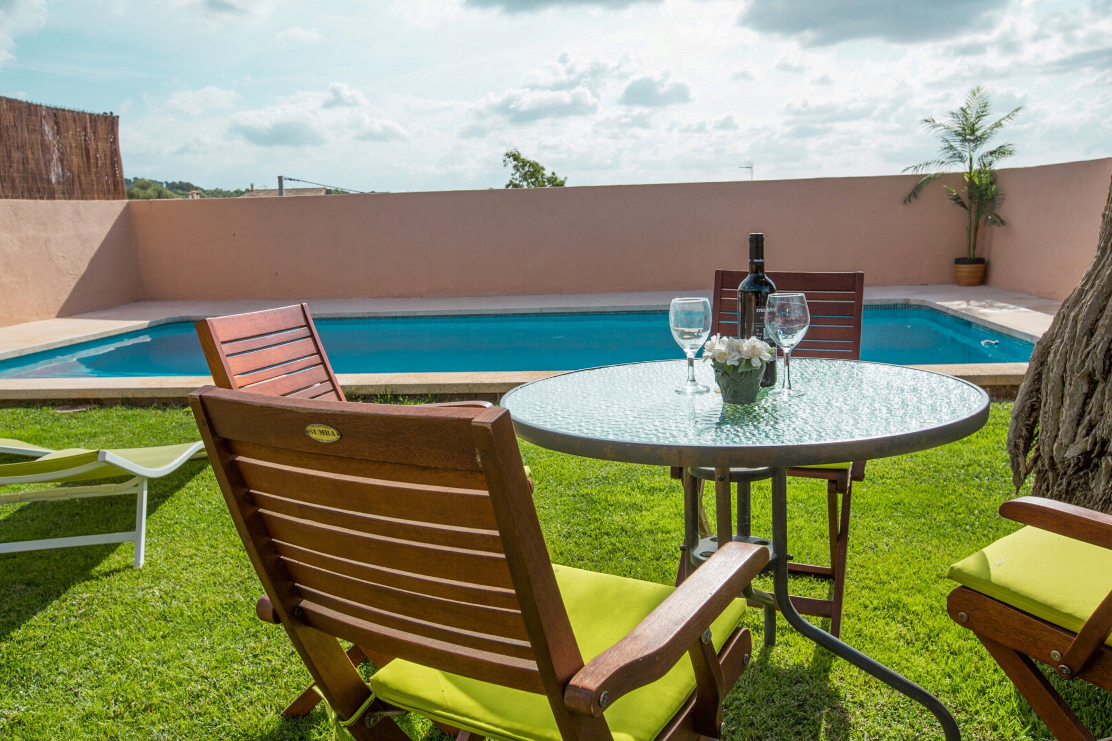 A poolside area with a round table, two wooden chairs with cushions, a bottle, and a glass on the table.