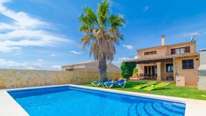 Piscina com espreguiçadeiras, uma palmeira e uma casa com parede de pedra e telhado de azulejos.