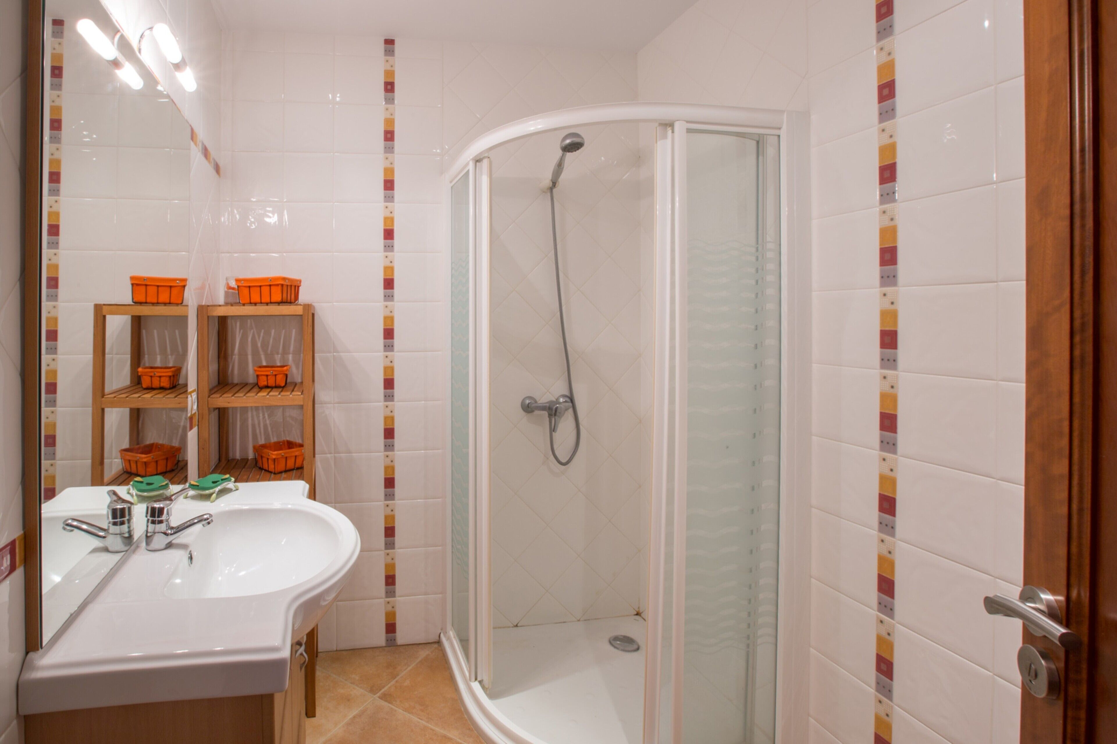 A bathroom with a white sink, mirror, and shower. There are orange baskets on a shelf and a wooden cabinet.