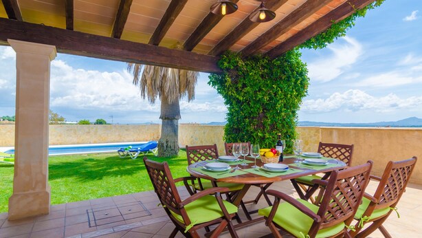 Um pátio ao ar livre com mesa de madeira posta para refeições, rodeado por cadeiras com almofadas verdes, vista para uma piscina e céu limpo.