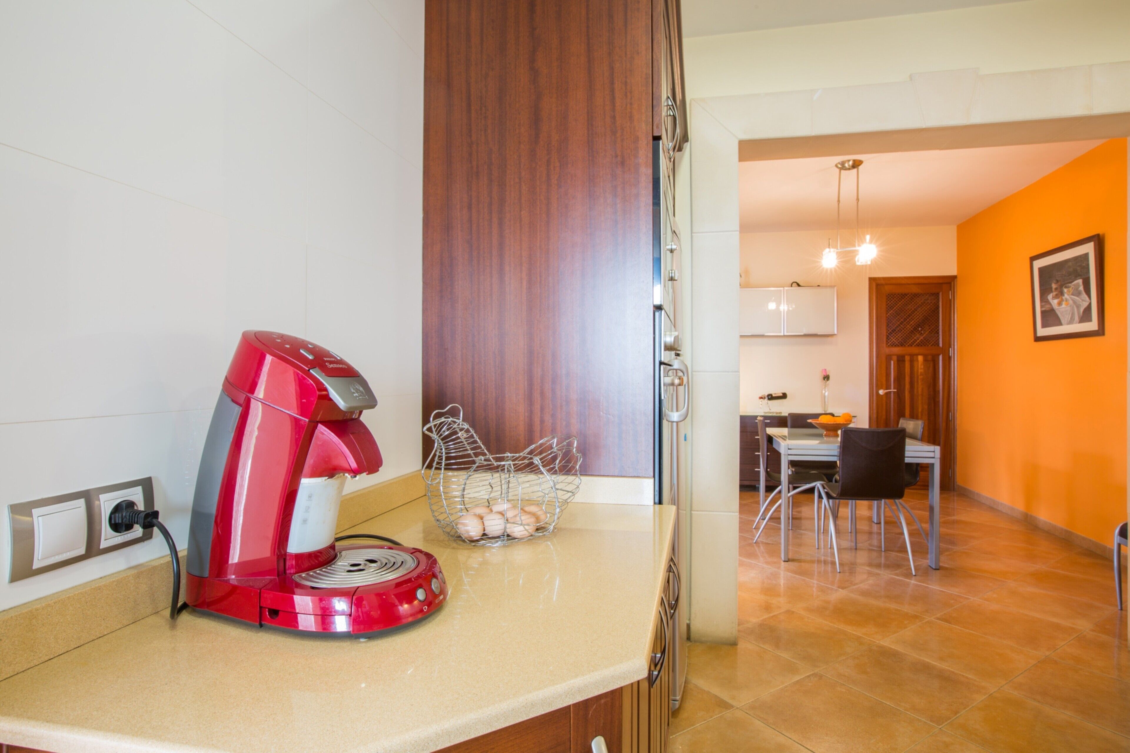 Una cocina con una cafetera roja, una cesta de huevos y un comedor con sillas y una mesa.