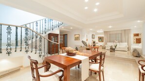 Ampla sala de estar com mesa de jantar de madeira, cadeiras, escada com grades ornamentadas e cozinha moderna ao fundo.