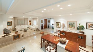 Uma sala de estar ampla com área de jantar, com uma grande mesa de madeira e cadeiras combinando, um sofá branco e uma lareira decorativa.