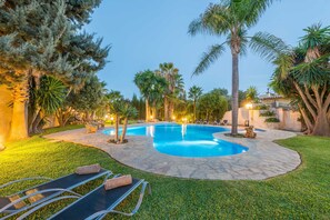 Une piscine à bord incurvé, entourée d’une terrasse en pierre et d’une végétation luxuriante, notamment des palmiers et une pelouse bien entretenue.