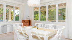 Un comedor con una mesa y sillas blancas, una lámpara colgante mullida de color blanco, y ventanas amplias con cortinas.