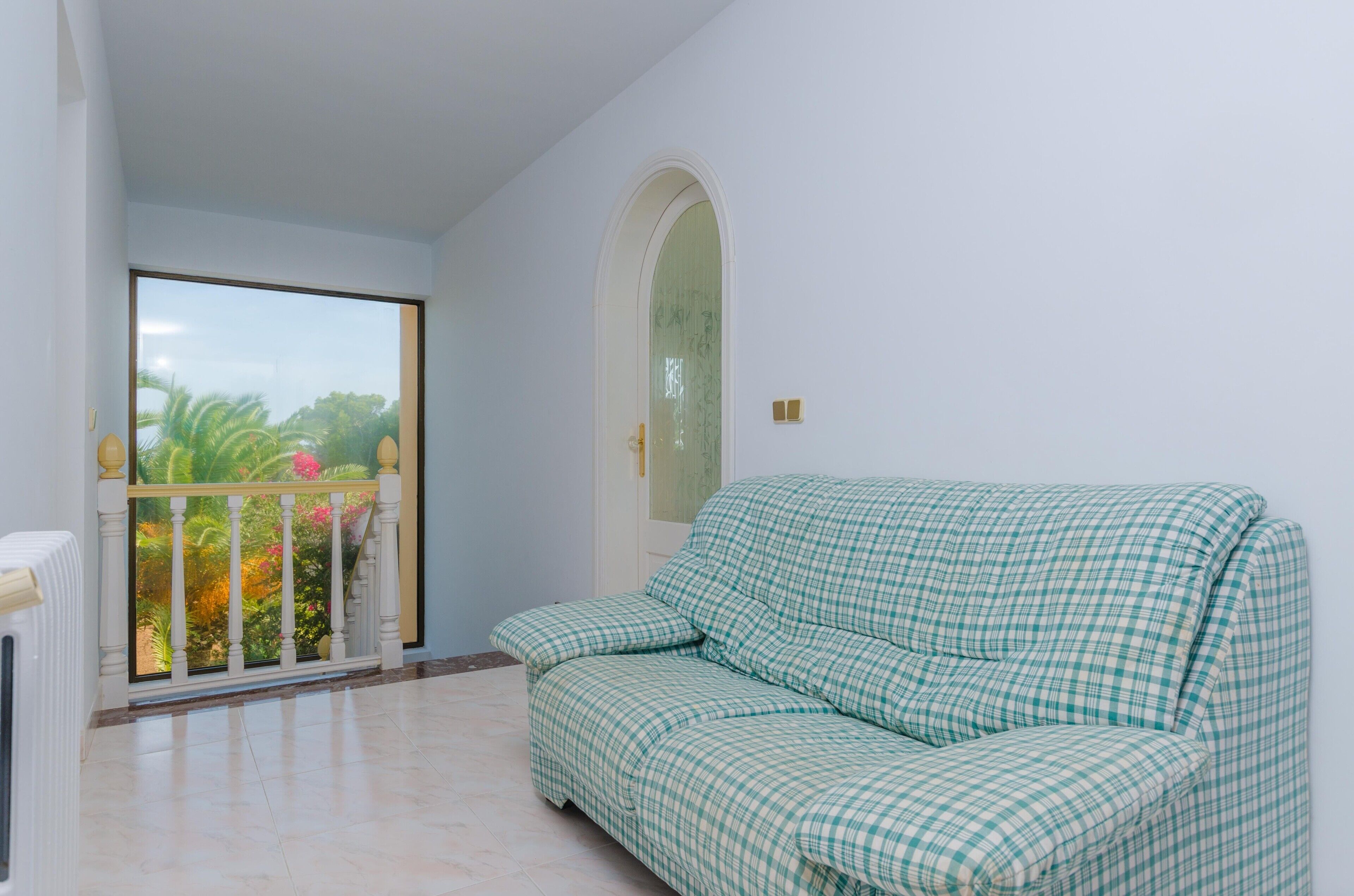 A room with a checkered sofa, a balcony with a view of greenery, and a white door with a window.