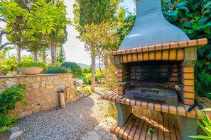 An outdoor brick oven grill with a stone wall and greenery in the background.