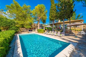 A rectangular swimming pool with clear blue water, surrounded by lounge chairs and umbrellas, with trees and a stone building in the background.