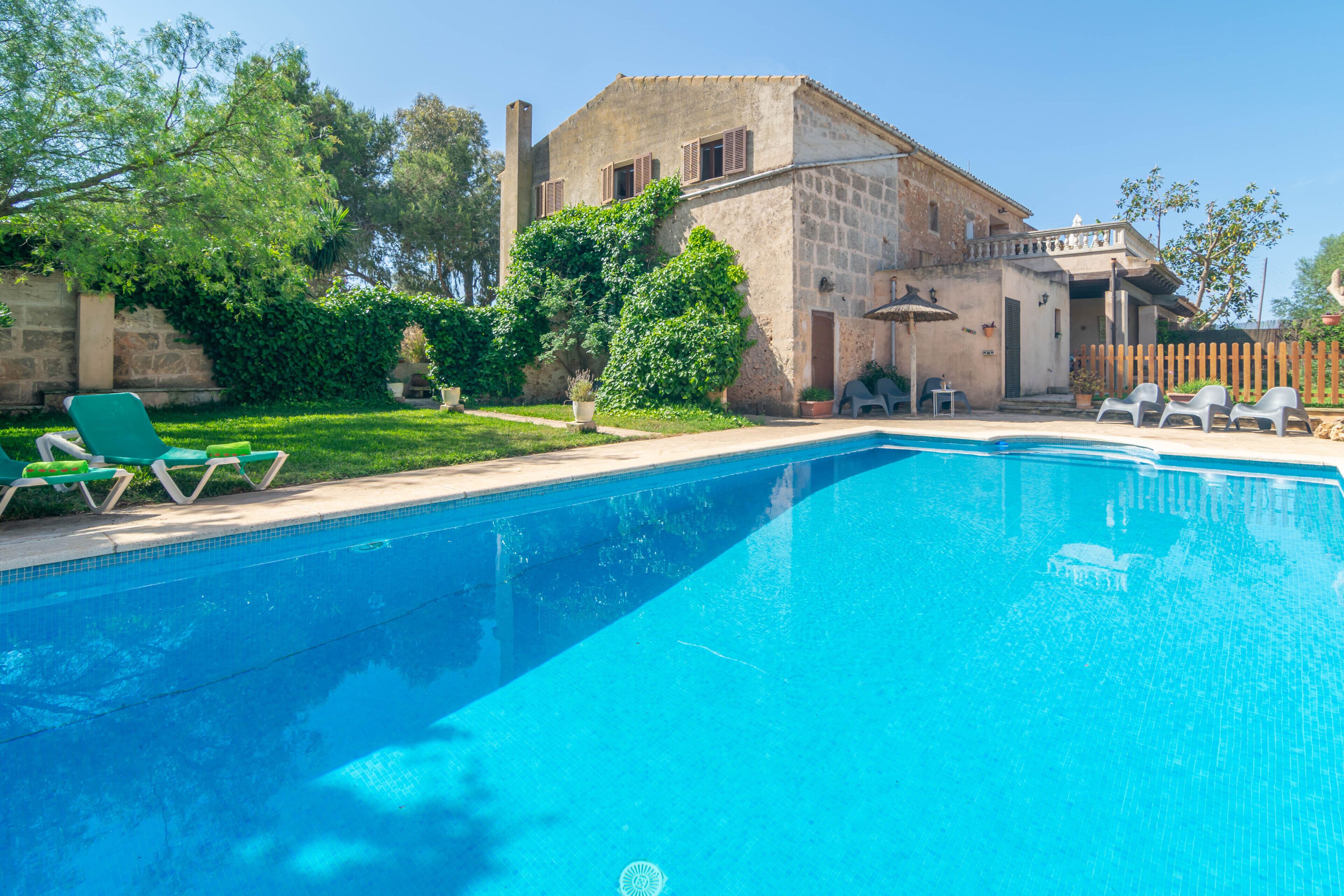 A swimming pool with lounge chairs, a stone building with shutters, and a wooden fence.