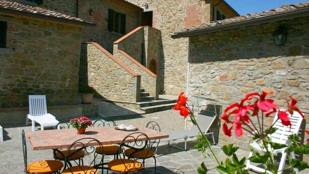 Un bâtiment en pierre avec un toit recouvert de tuiles, un escalier, un espace extérieur aménagé avec une table et des chaises, et des plantes en pot.