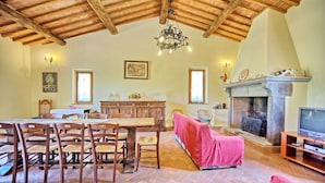A spacious living room with a high wooden ceiling, a fireplace, a dining area with wooden furniture, and a television.