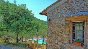A stone building with a window and a door, surrounded by trees and a fence.