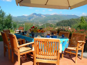 Un área de comedor al aire libre con una mesa dispuesta para cuatro personas, rodeada de sillas de madera, con vistas a un paisaje montañoso.