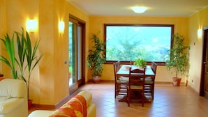 Un espace repas comprenant une table et des chaises, entouré de plantes en pot, et de grandes fenêtres donnant sur un paysage verdoyant.