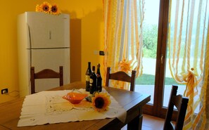 Una mesa de comedor con mantel blanco, sillas de madera y un ramo de girasoles. Hay botellas de vino sobre la mesa, y se ve un refrigerador al fondo.