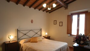Quarto com cama, duas luminárias de cabeceira, ventoinha no teto, janela com persianas e mesa de cabeceira com um vaso de flores.