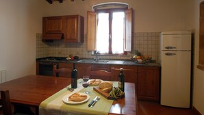 Cozinha com armários de madeira, uma janela com cortinas, uma geladeira e uma mesa de jantar posta com pão, vinho e uma garrafa.