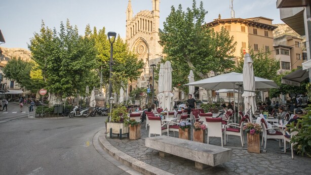 Un café al aire libre con mesas y sillas, rodeado de árboles y un edificio histórico al fondo.