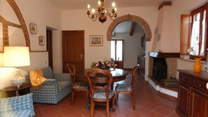 A dining area with a round table, chairs, and a chandelier. There is a fireplace and a sofa with a lamp. - Beautiful apartment for 4 guests with WIFI, A/C, pool, TV and pets allowed, close to San Gimignano (Montaione)