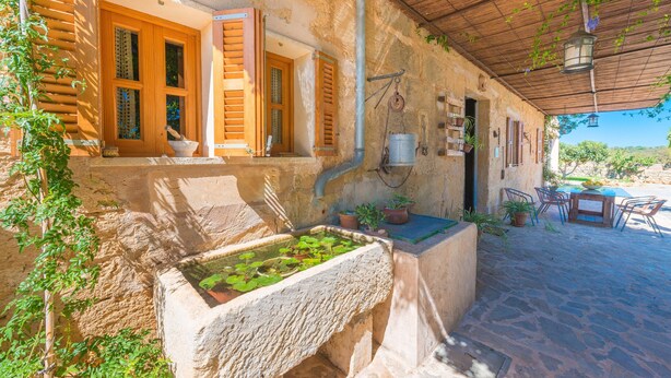 Un edificio rústico de piedra con contraventanas de madera, una fuente de piedra y asientos al aire libre.