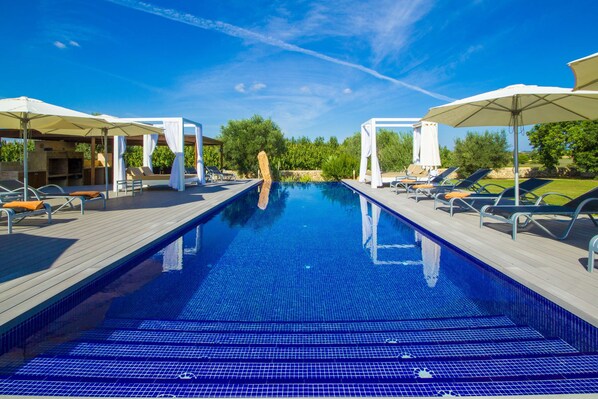 A swimming pool with sun loungers and umbrellas, surrounded by greenery and a clear sky.