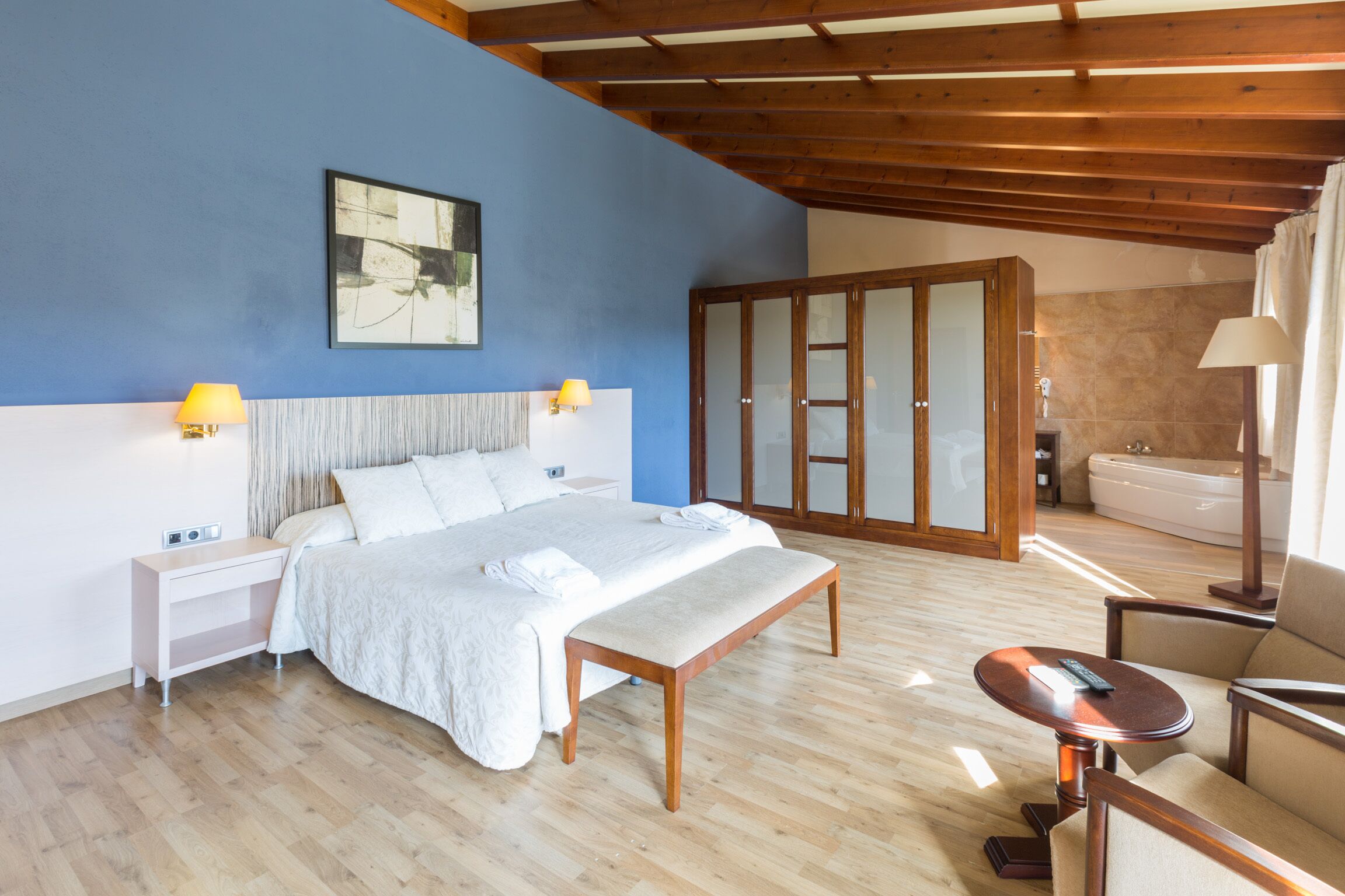 A bedroom with a bed, bedside tables, a bench, a chair, a wooden ceiling, and a bathroom with a bathtub.