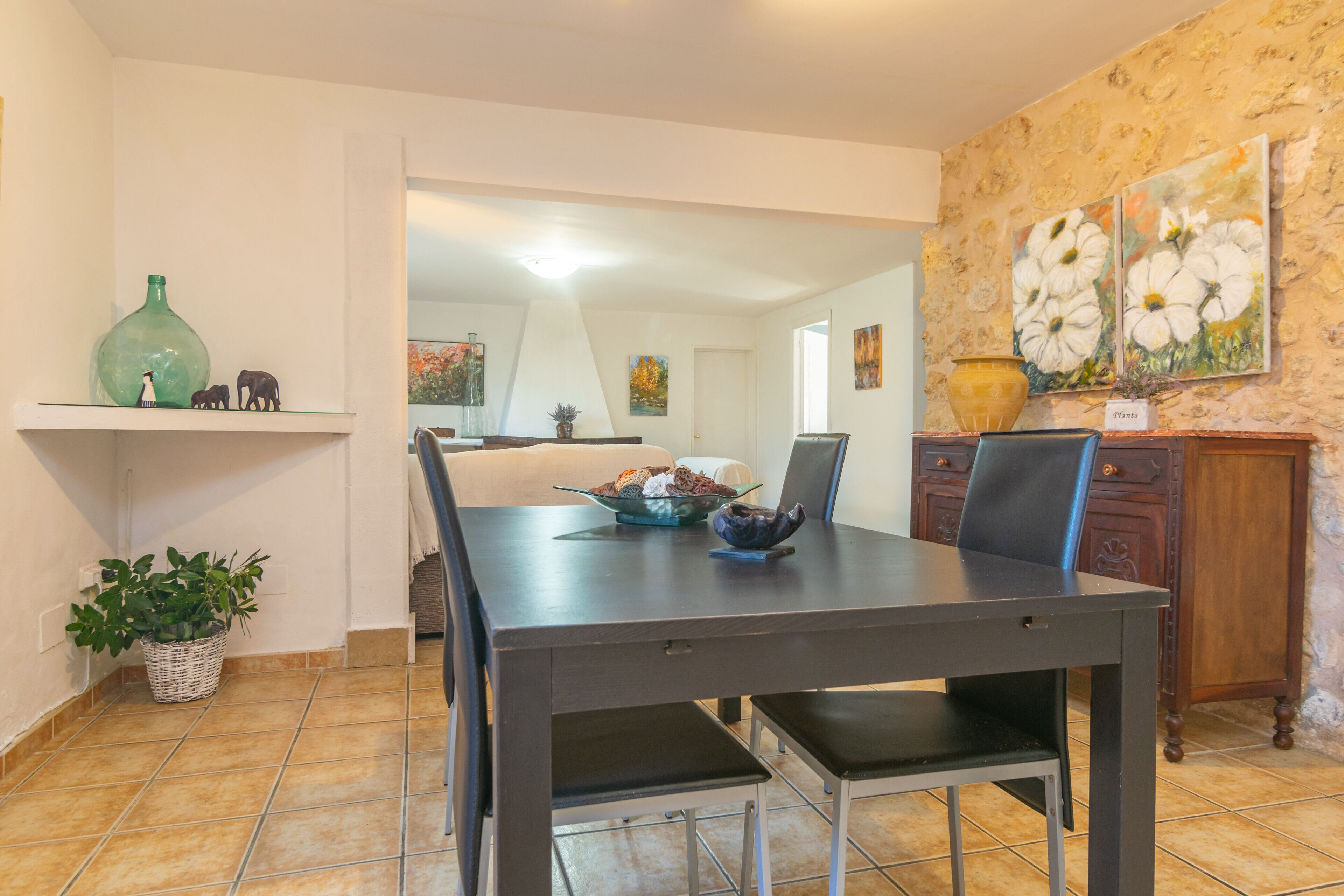 A dining area with a dark wooden table, black chairs, and a bowl of fruit. There is a potted plant on the floor and a vase on the wall.