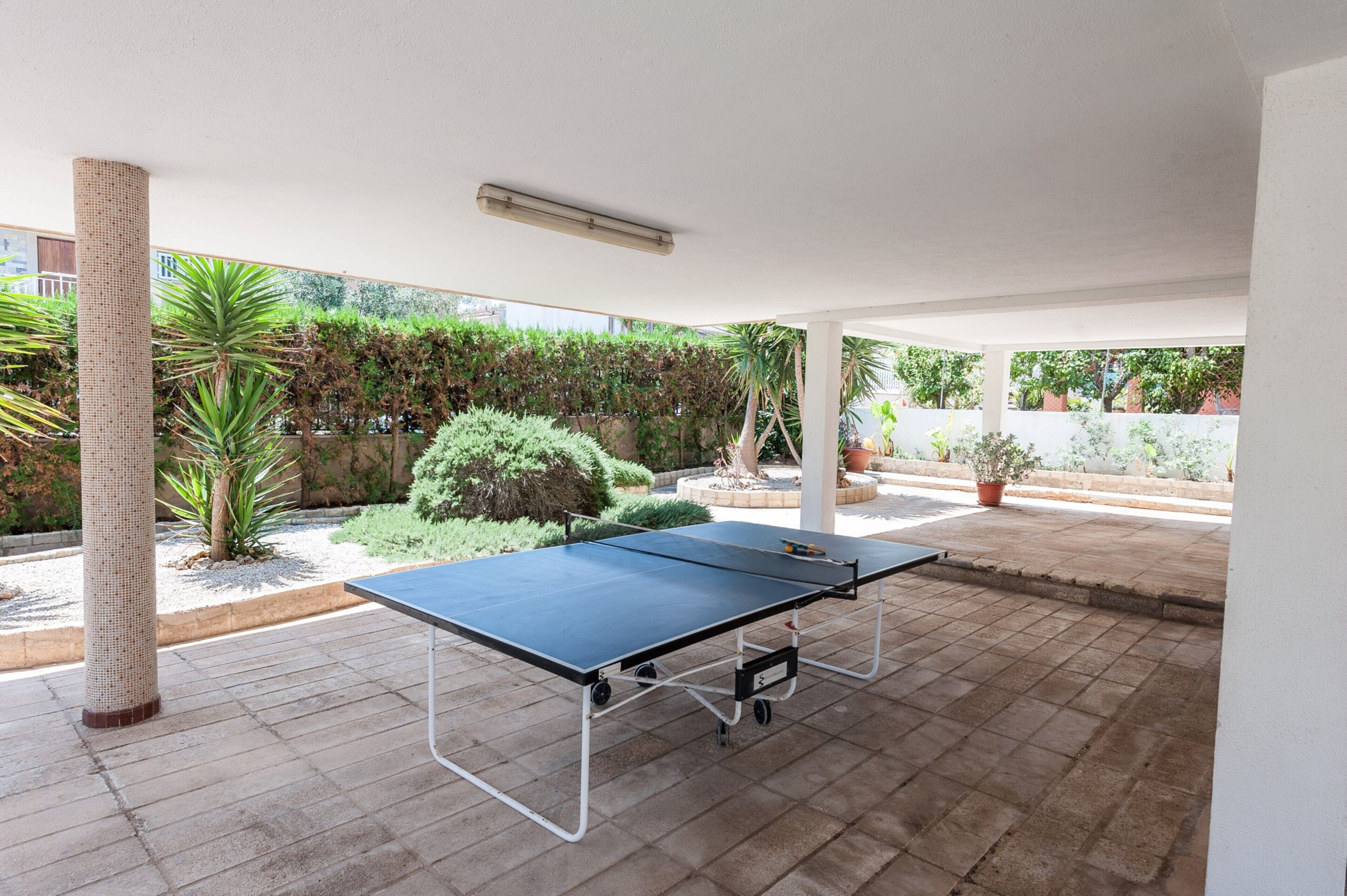 Ein Tischtennis-Tisch in einem überdachten Außenbereich mit Ziegelboden, umgeben von Blumentöpfen und Blick auf einen Garten.