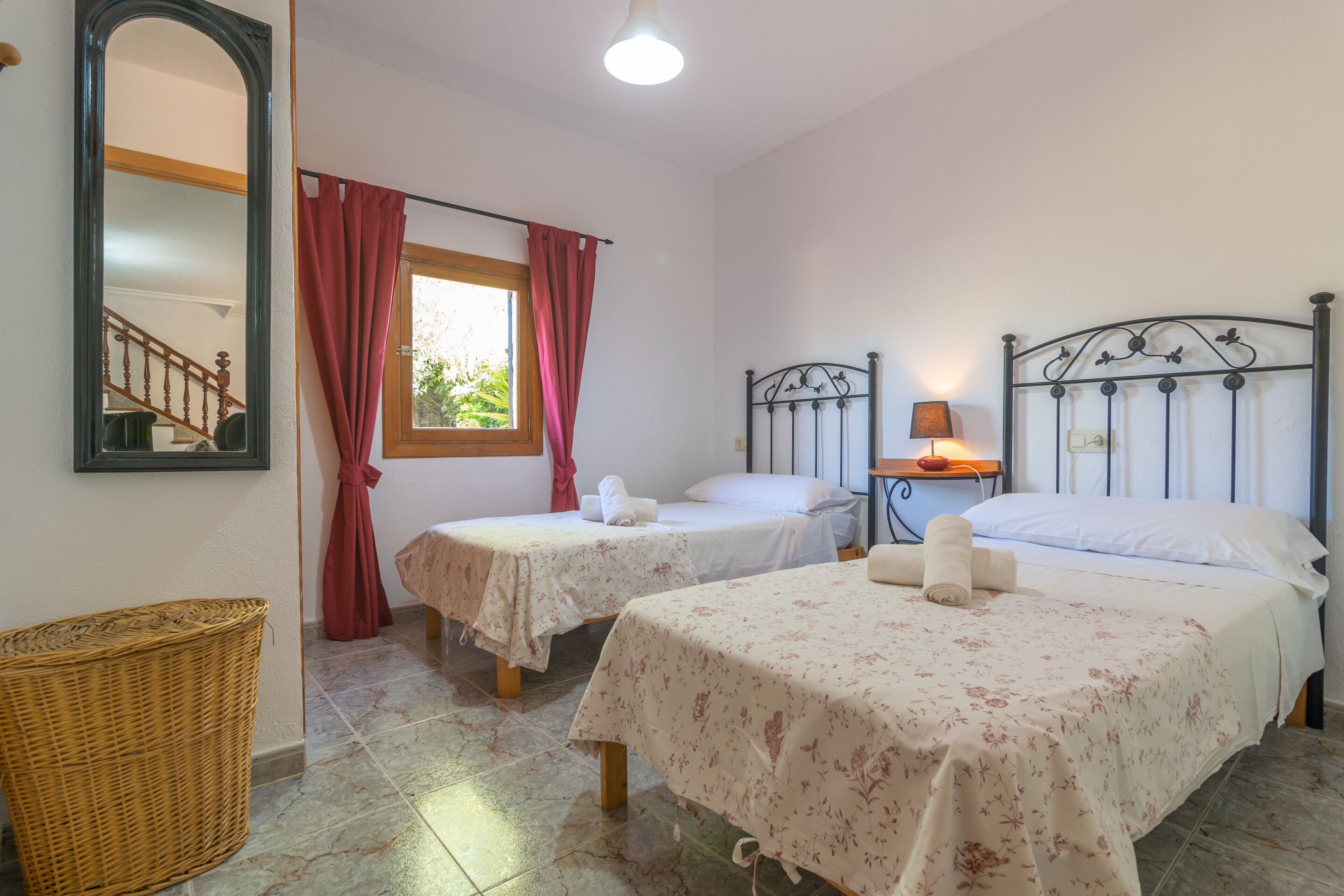 A hotel room with two single beds, a large mirror, a window with red curtains, a wicker basket, and a lamp on a bedside table.
