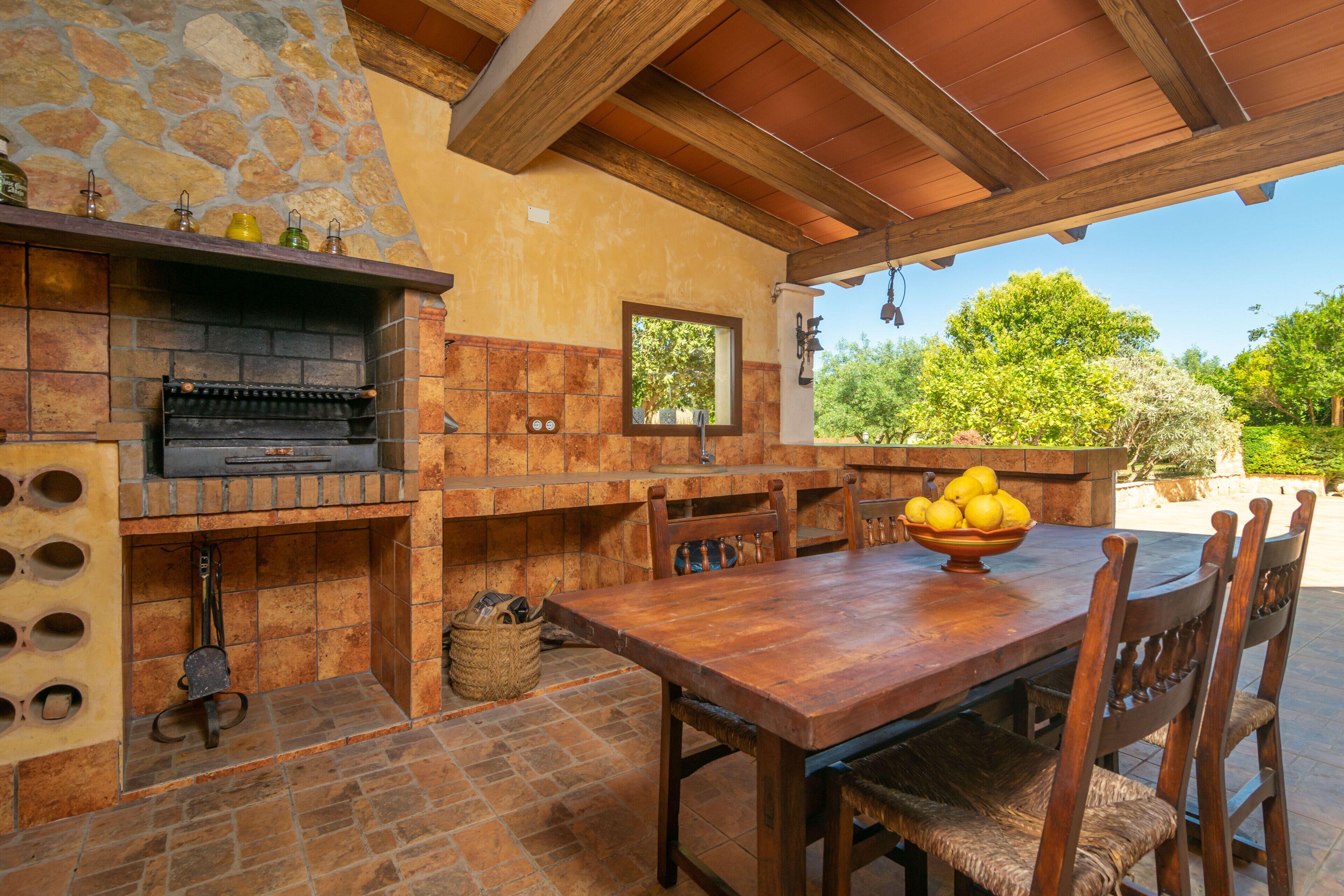 Une cuisine extérieure rustique, avec un mur en pierre, des poutres en bois et une table en bois accompagnée de chaises.
