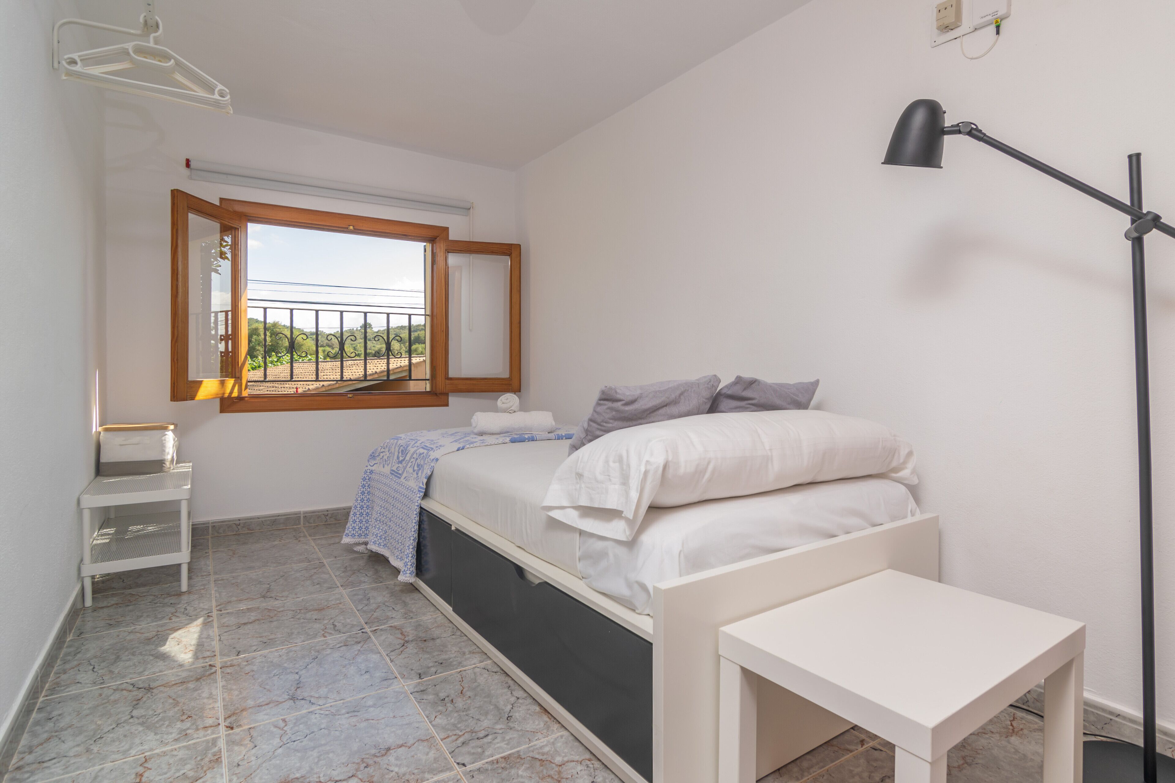 A bedroom with a bed, a bedside table, a floor lamp, a window, and a small white shelf.
