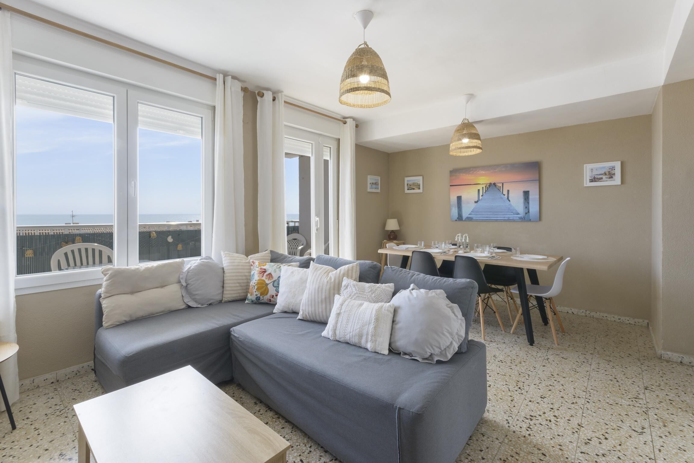 Un salón con un sofá gris, una mesa de comedor y un cuadro de una puesta de sol.