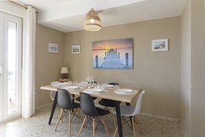 Un coin repas avec une table en bois dressée pour quatre personnes, des chaises noires et blanches, un luminaire suspendu et un tableau représentant un coucher de soleil accroché au mur.