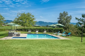 Une piscine entourée de chaises longues et de parasols, elle-même bordée d’une pelouse bien entretenue et d’arbres, avec des montagnes en toile de fond.