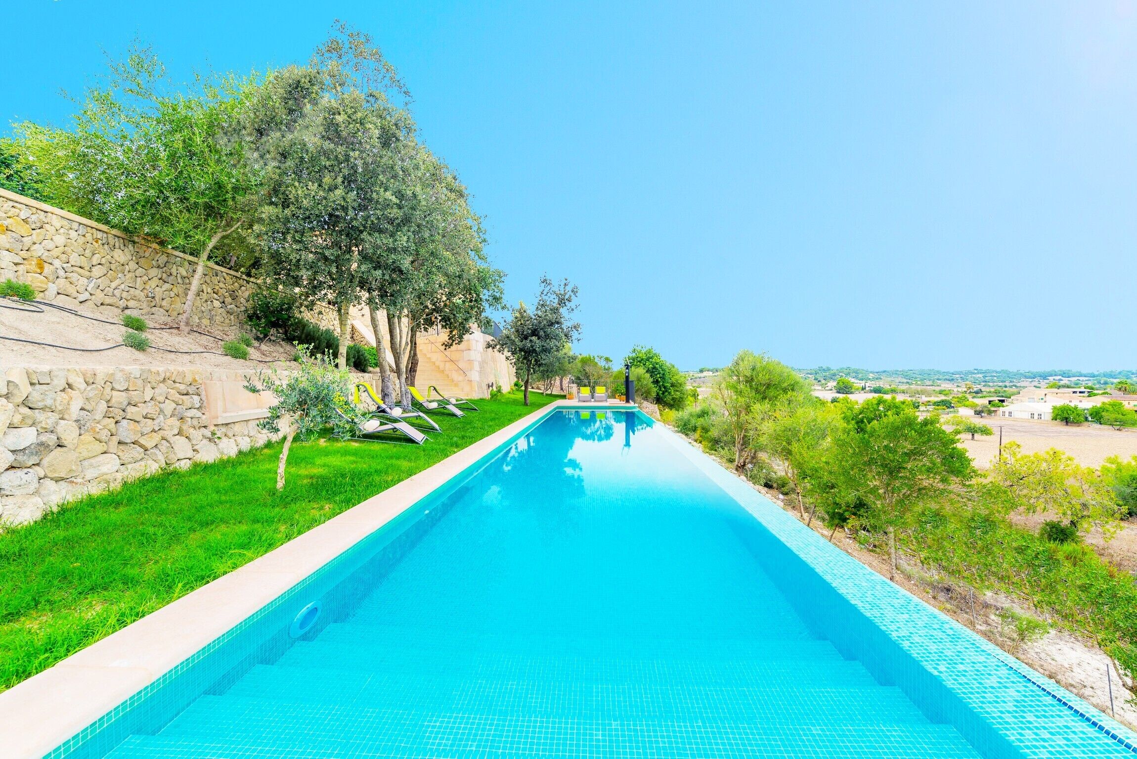 Une piscine rectangulaire à l’eau d’un bleu limpide, entourée d’un mur de pierre et d’une végétation luxuriante.