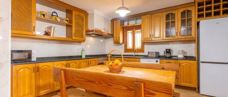 A kitchen with wooden cabinets, a marble countertop, a wooden dining table with a bench, and a white refrigerator.