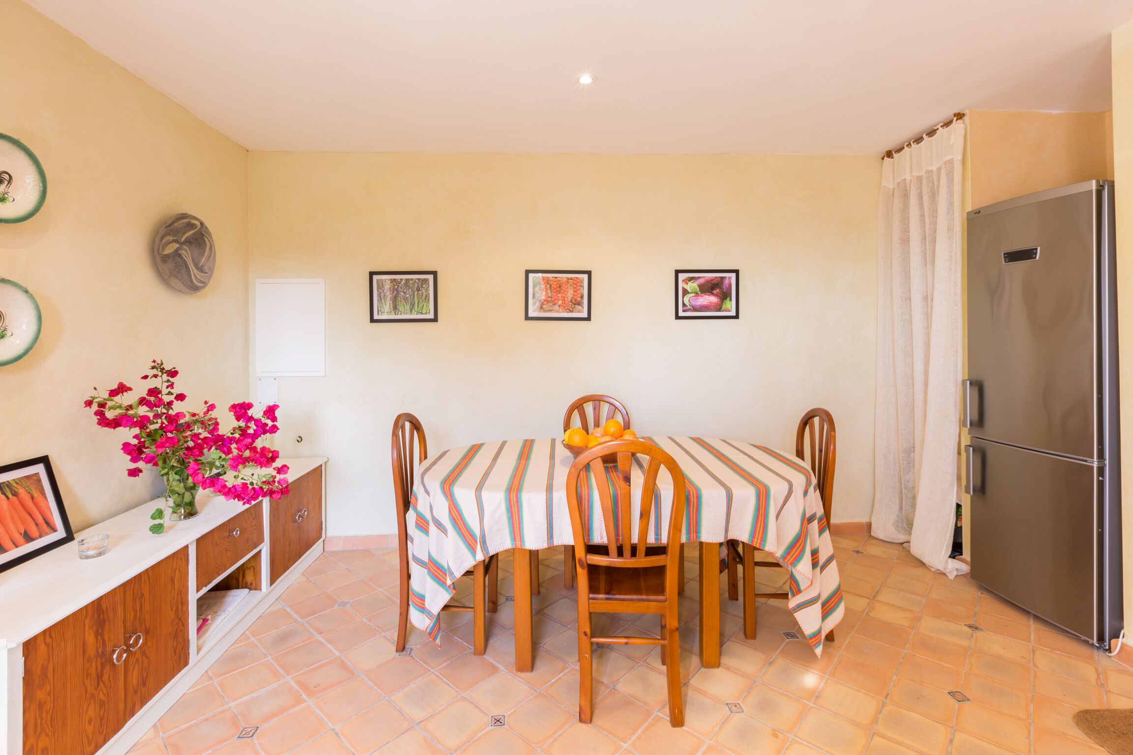 A dining area with a table set for four, surrounded by chairs, a refrigerator, and a cabinet with decorative items.