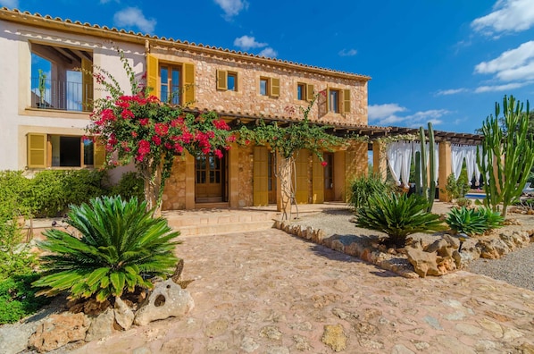 A two-story stone building with a tiled roof, a courtyard with a cobblestone path, and various plants including a prominent pink flowering bush.