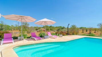 A swimming pool with lounge chairs and umbrellas, surrounded by greenery and a clear sky.
