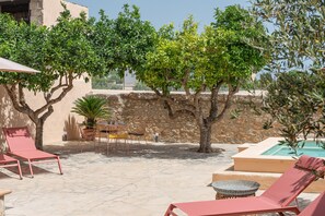 An outdoor patio with stone flooring, red lounge chairs, a small table with chairs, and a pool area.