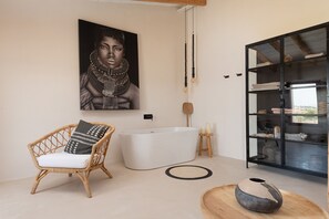 A modern bathroom with a freestanding bathtub, a wicker chair with a cushion, a large framed artwork on the wall, and a glass cabinet with towels.
