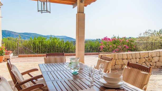 An outdoor patio with a wooden table set for dining, surrounded by chairs, and overlooking a scenic landscape with mountains and a stone wall. - VILLA BELLAVISTA – Villa with private pool in Campanet with free WiFi (Campanet)