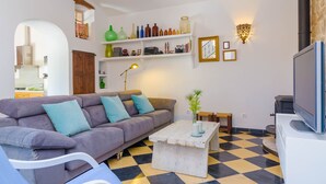 Un salon comprenant un canapé, une table basse et un téléviseur. On y trouve également des étagères avec divers objets et une lampe.