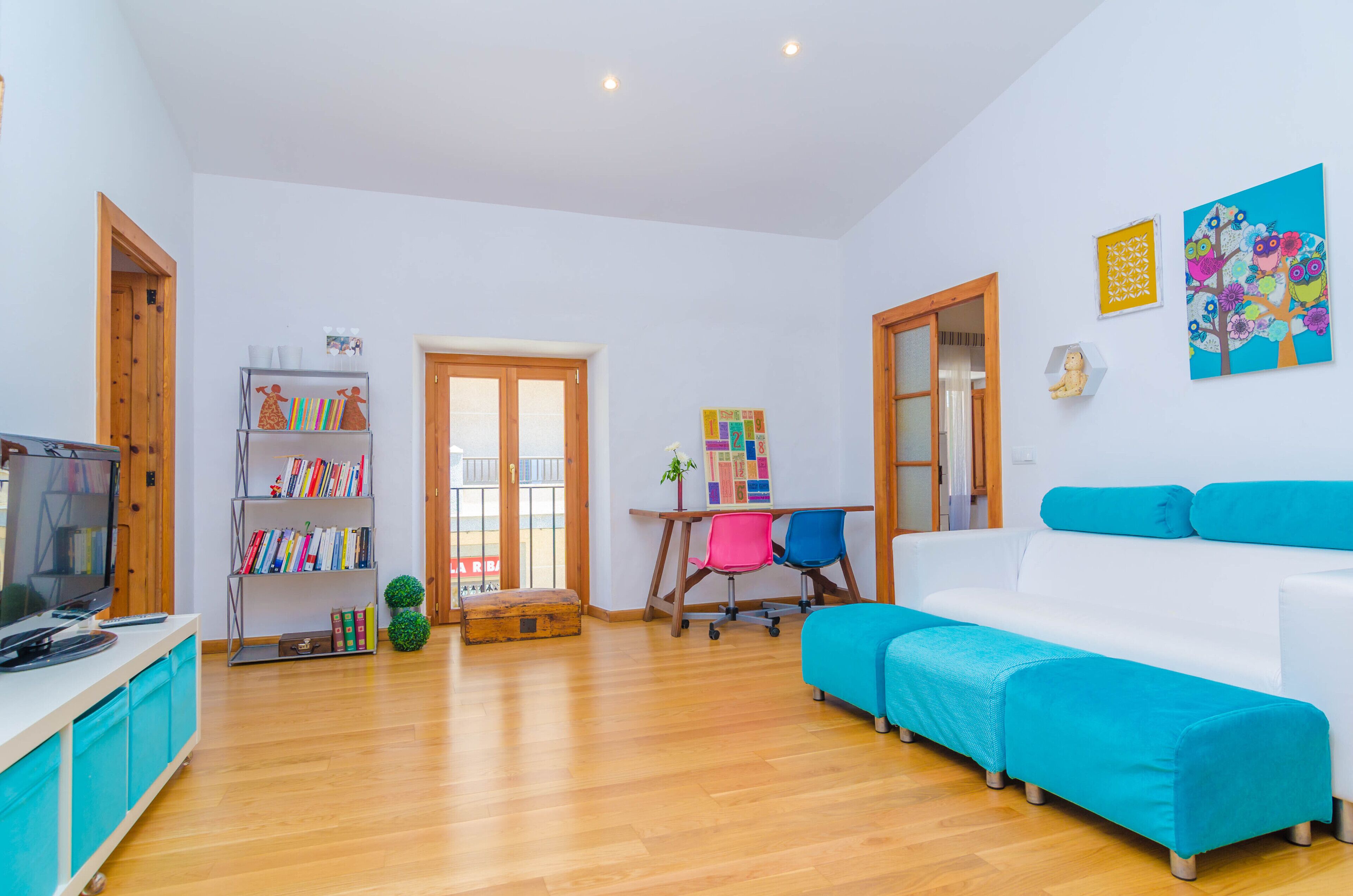 A modern living room with a white sofa, wooden flooring, a television, a bookshelf, and a colorful painting on the wall.
