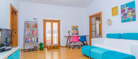 A modern living room with a white sofa, wooden flooring, a television, a bookshelf, and a colorful painting on the wall.
