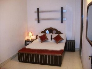 A neatly made bed with a wooden headboard, a bedside lamp, and a small bedside table. - Anmol Hotels (New Delhi)